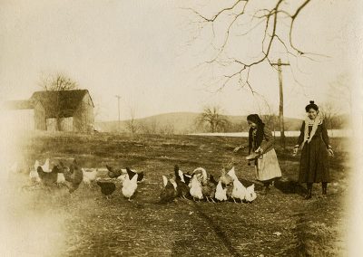 Black and white photo from early 1900s of 2 Native girls feeding chickens. One girl is recognized as Annie Coodlalook.