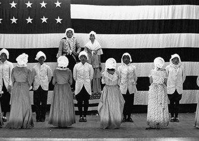 Black and white photo of Native children in colonial costumes on a stage in front of a large American flag