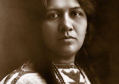 1900 black and white portrait of Angel de Cora, a young Native woman with bradied hear wearing a traditional beaded dress