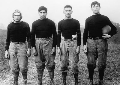 1912 black and white photo of 4 Native youth in early 20th C football uniforms posing for the camera. Jim Thorpe stands at far right holding a football.