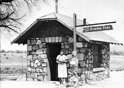 1936 black and white photo of Wa-Pai-Shone traditing post- a small rough stone building with a woman posing in front