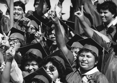 1984 candid black and white photo of Native youth in graduation caps and gowns smiling and holding their right arms in the air with their pointer fingers raised.