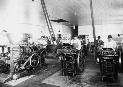 1920 black and white photo of a printing press room with young Native men standing over and working at printing machines.