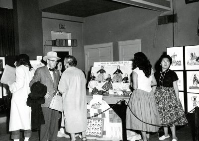 1950 black and white photo of handmade cloth dolls on display at an art event. People mill around looking at the art and talking to each other.