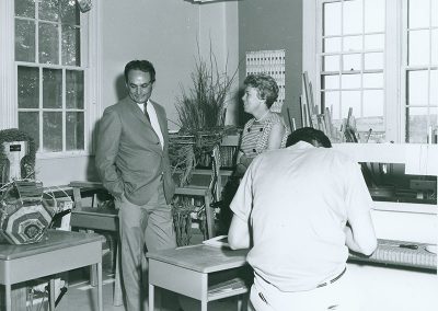 1970 black and white photo of Lloyd Kiva New in a suit standing in weaving classroom with a woman with short blond hair. A young man is hunched over a loom with his back to the camera in the foreground.