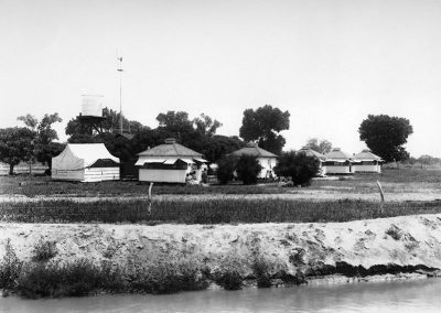 Black and white photograph of land across a body of water with square wooden structures and sparse trees.