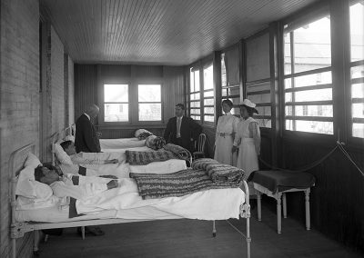 Black and white photo of a turn of the century infirmary. Two doctors and 2 nurses stand by beds with young men.