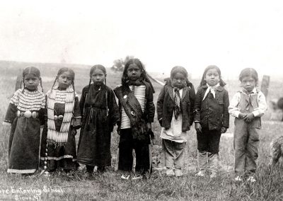 Black and white photo of American Indian children lined up in front of the camera in native dress. Late 19th C.