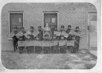 1900 black and white photo of young boys in military style uniforms and caps posing with an older woman in front of a brick building. All the boys hold American flags.