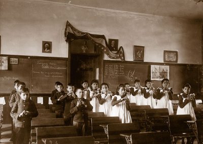 Black and white late 19th C. photo of Native children in uniform standing by their desks making gestures with their hands.