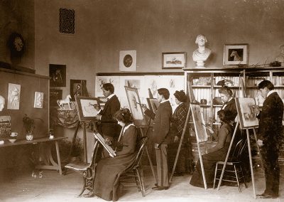 1900 black and white photo of young Native men and women at easels in a drawing still life objects
