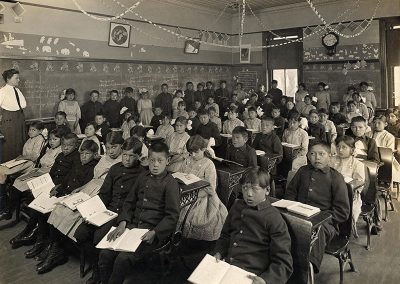 1911 black and white photo of Native children in uniforms seated in classroom with open books