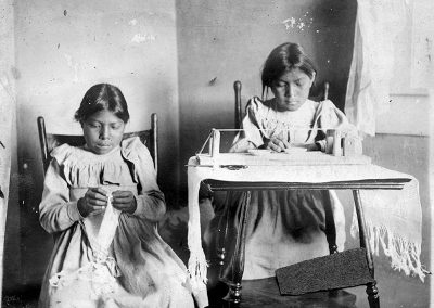 1905 black and white photo of 2 young Native girls working on beading. One girl sits at a desk with a traditional bead loom and the other sits beside with indistinct fabric in her hand.