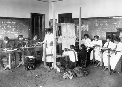 Early 1900s black and white photo of Native youth in an art class weaving and working with fibers