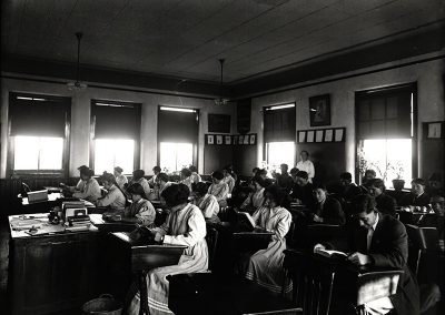 1913 black and white photo of older Native students in a classroom reading at their school desks.