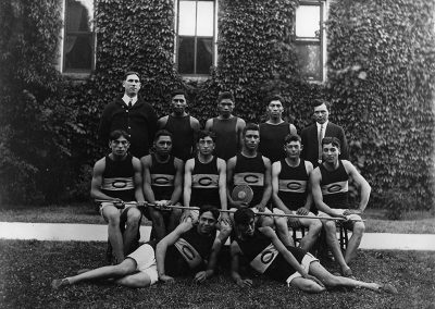 1913 black and white photo of team of young Native men in track and field style shorts and sleeveless shirts posing on lawn in front of an ivy covered building with adult males on either side in the back row.