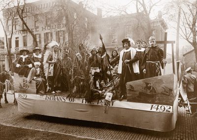 1920 black and white photo of parade float titled "Indians of the Past" showing Native youth in traditional dress, Puritan costumes and Columbus with a priest