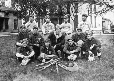 1913 black and white photo of Native youth baseball team with adult male in center. They stand on a grassy lawn with several buildings behind them.