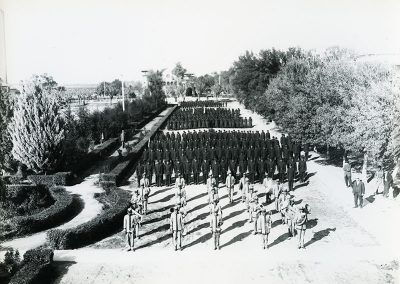 1900 black and white aerial photo of uniformed men and women in formation. A marching band stands in the foreground and the rest of the individuals are in dark uniforms in formation behind them.