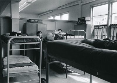 1970 black and white photo of large dorm hall with multiple metal framed bunk beds with 2 young men. One is lounging on an upper bunk reading and the other stands next to the bed.