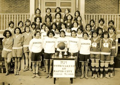 1929 black and white photo of large group of young Native women from 4 different basketball teams. They pose for the camera in different uniforms with trophies on a table and a banner saying "1929 Tournament at Rapid City Indian School" in the foreground.