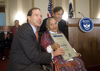 2006 color photo of an elderly Native woman in a wheelchair posing with 2 Caucasian men. She is holding a plack with her name "Ester Martinez" and underneath "Master Artist." They are all smiling.
