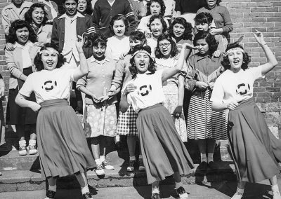 1950 black and white photo fo 3 very animated Native cheerleaders in front of a crowd of students on steps to a building in the background.