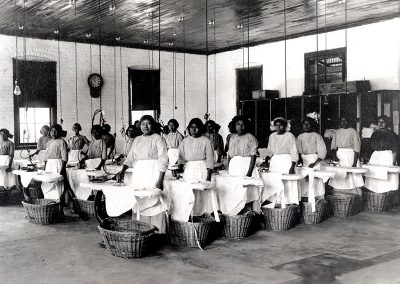 1910 black and white photo of young Native women in a large industrial style laundry room at ironing boards, baskets below.