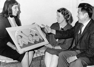 1963 black and white photo of young Native woman happily showing her painting to a Native man and Caucasian woman.
