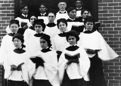 1902 black and white photo of young Native women and men in ecclesiastical choir outfits holding hymnals posed on steps at arched building entrance. An older Caucasian male clergy stands with them at the back.