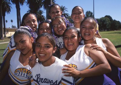 1999 candid color photo of young Native women in cheeleading outfits grouped tightly together and laughing and smiling for the camera