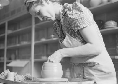 1940s black and white photo of young woman shaping a vessel on a pottery wheel with shelves of pottery behind her.