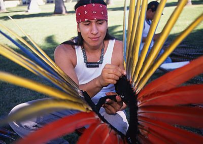 1999 candid color photo of a young Native man wearing a red bandana around his forehead and carefully aligning long feathers on a head dress