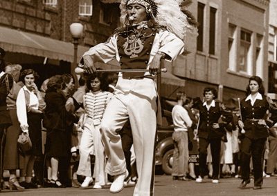 1940 black and white photo of a male drum major in marching costume with headress carrying a twirling batton in both hands and a whistle in his mouth. He marches down the middle of a street in front of the rest of the band with bystanders looking on.