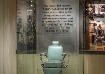 Old style barber chair with metal arms and smooth teal blue leather like materal enclosed in a large glass case. Black braids and cut hair lay around the chair on the floor.