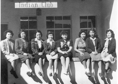 1930s black and white photo of young Native women in dresses and blazers sitting on a stucco wall posing for the camera in front of a veranda with a sign saying "Indian Club"