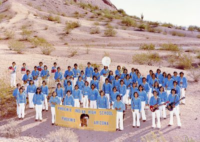 1982 color photo of Native youth band members in blue velvet tunics tied at the waist with concho belts and white trousers. All are holding their instruments and a yellow banner which says "Phoenix Indian High School, Phoenix, Arizona." They stand in a desert setting.
