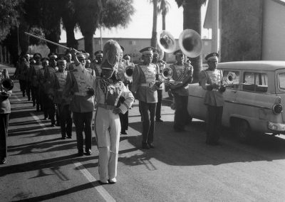 1960 black and white photo of a marching band on the road with a drum major in front with her baton raised and whistle in her mouth.